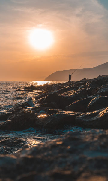 fotografia di un tramonto sul mare, sullo background si vede la sagoma di un uomo che pesca